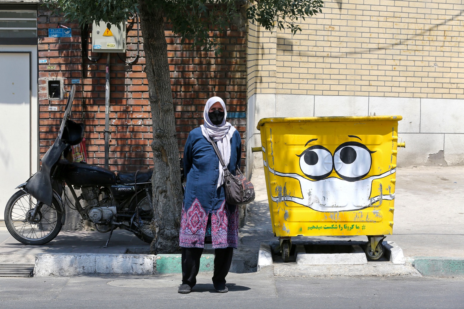 Foto Atta Kenare/AFP Em um bairro no sul de Teerã, capital do Irã, uma mulher passa ao lado de uma caixa de coleta de lixo pintada com uma máscara de proteção contra a covid-19. Foto Atta Kenare/AFP