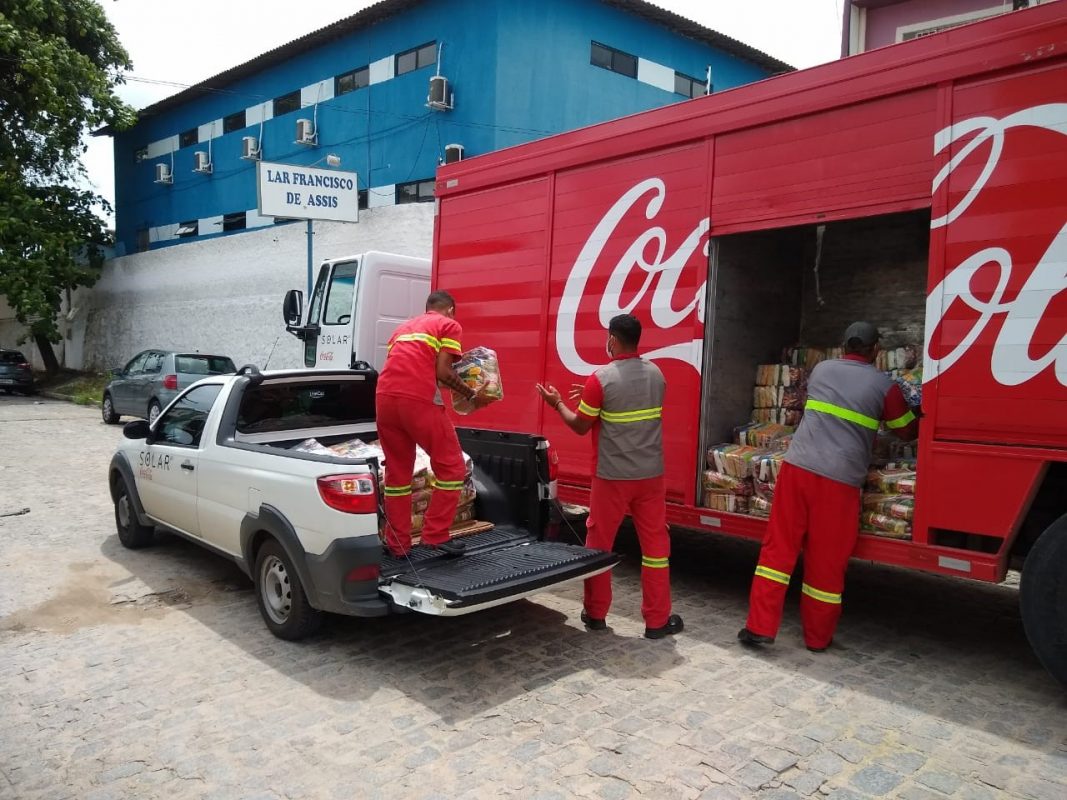 Três funcionários uniformizados da Coca-Cola Brasil transportam cestas básicas, movendo-as de uma picape branca, posicionada à esquerda, para o caminhão vermelho da Coca-Cola que está no canto direito.