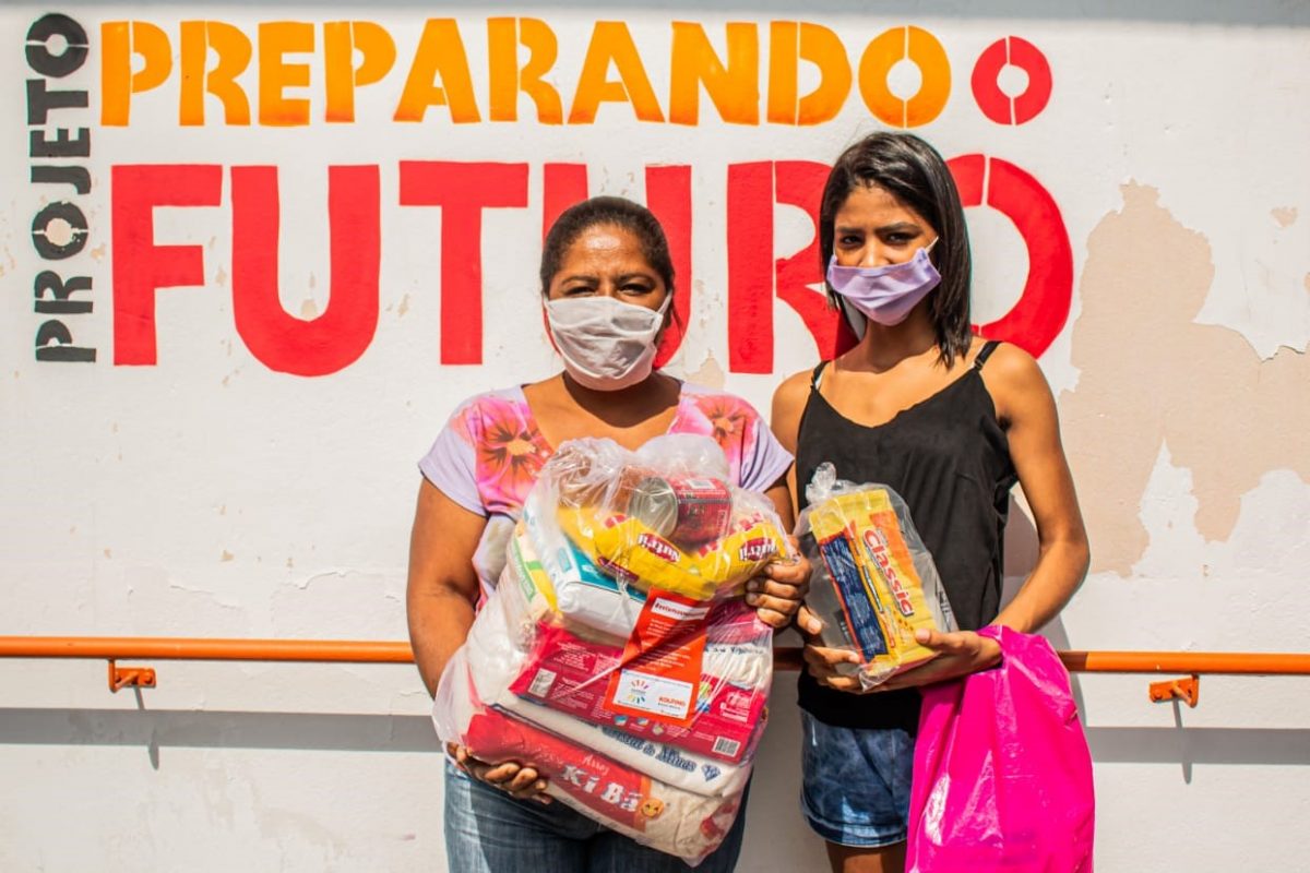 Uma senhora e uma adolescente posam para uma foto, segurando cestas básicas. A senhora, à esquerda, usa calça jeans e uma camisa florida, com uma máscara branca, enquanto a jovem à direita usa uma camiseta preta, short jeans e uma máscara roxa. Atrás delas, pode-se ler na parede "Projeto Preparando o Futuro"
