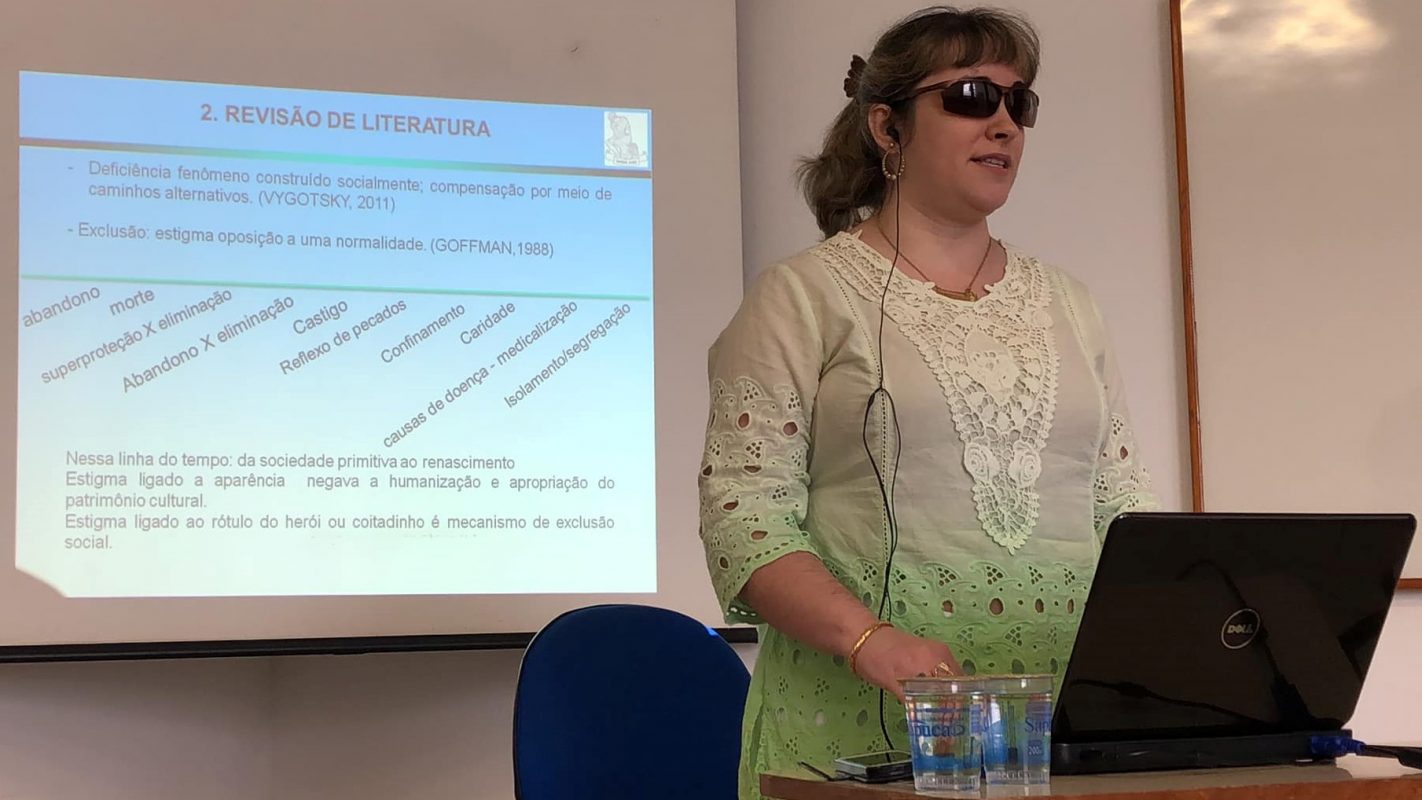 Luciane Molina é uma pessoa com deficiência visual especialista em acessibilidade. Ela está de pé, de óculos escuros, em frente a um notebook. Há uma slide sobre uma aula projetado na tela atrás dela e dois copos de água mineral na mesa. à frente. Luciane está com o cabelo preso em um rabo de cavalo