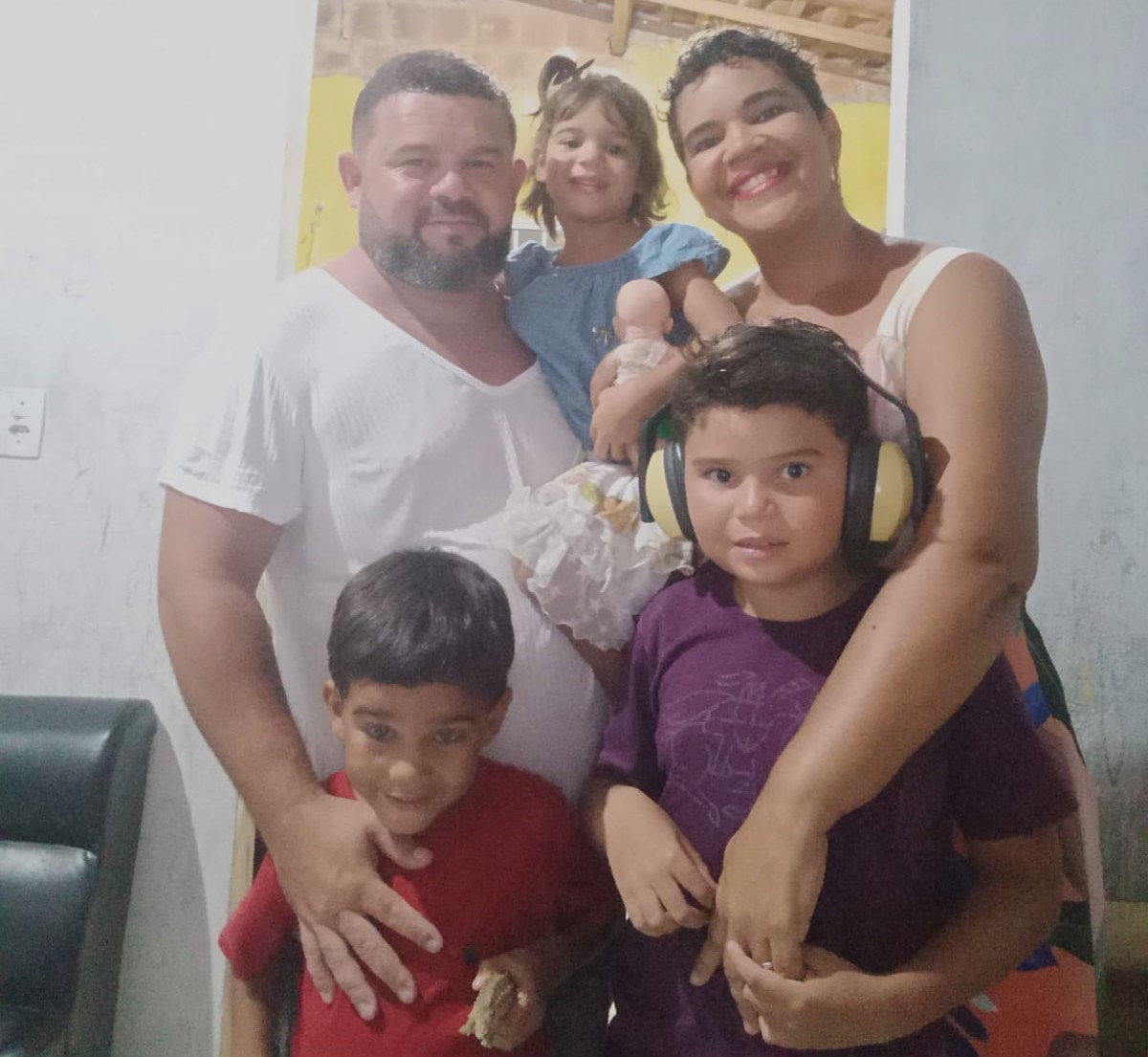 Foto colorida de Lucielys, seu esposo Ibson Magalhães Lucas e seus filhos. Ibson, de barba e cabelo preto, usa uma camiseta branca e está com sua filha Iana, de vestido azul, no colo. Lucielys está ao lado dele, abraçada em Ibrahim Elias, que está com uma blusa roxa e headfones, e ao lado dele João Raviv aparece com uma camiseta vermelha.