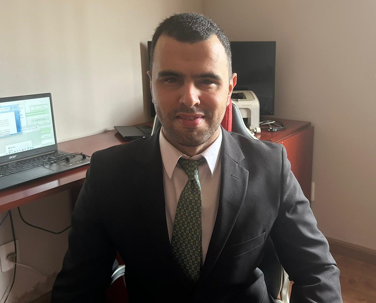 Foto colorida de Gabriel Henrique. Gabriel é cego, tem pele branca e cabelos pretos bem curtos. Ele usa um terno preto e uma gravata verde clara. Ao fundo, escritório e computador de mesa