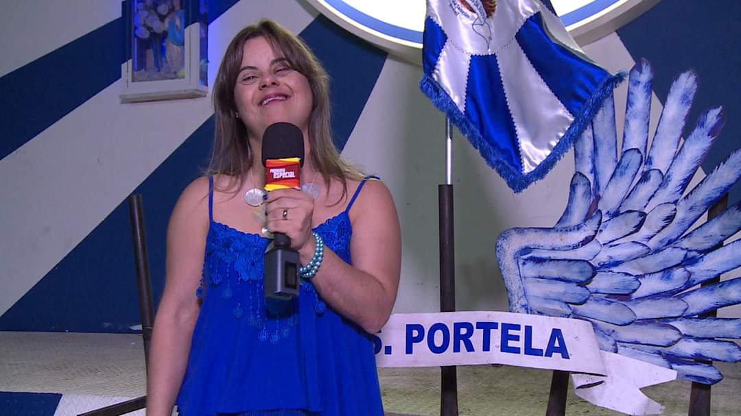 Foto colorida de Fernanda Honorato em gravação de reportagem na quadra da Portela. Fernanda é branca com cabelos loiros. Ela segura um microfone com a mão esquerda. Ela usa uma blusa azul e sorri para a câmera. Ao seu lado direito aparece uma bandeira da Portela e ao fundo decoração com as cores azul e branco