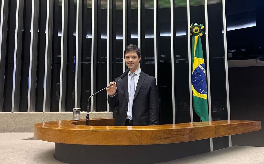 Foto colorida de João Vítor na Câmara dos Deputados em sessão pelo Dia Nacional de Luta da Pessoa com Deficiência. João é branco com cabelos pretos. Ele está de pé e usa um terno, ele segura o microfone da tribuna com a mão direita e sorri olhando para frente. A esquerda de João, bandeira do Brasil.
