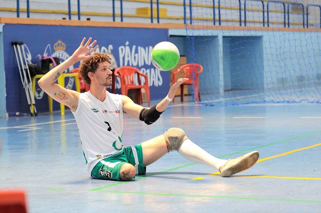 Foto colorida de Beto Maia jogando vôlei. Beto é um homem branco com cabelos castanhos e cacheados, barba preta. Ele está sentado e suas mãos levantadas, com a direita em posição de acertar uma bola de vôlei que está na sua frente. Ele possui uma das pernas amputadas e usa um short verde e uma camiseta branca. Ao fundo, ginásio de vôlei pintado de azul.
