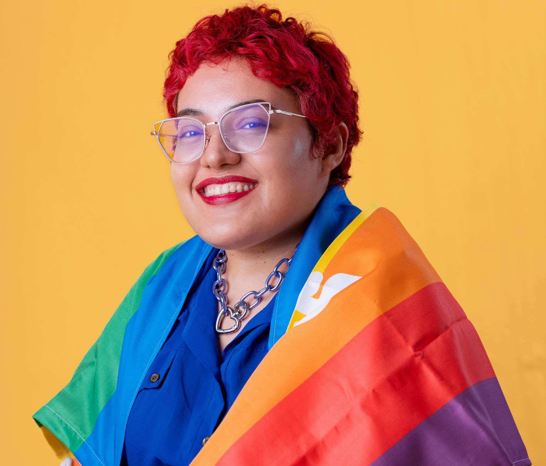 Foto colorida de Priscila Siqueira. Ela é uma mulher com nanismo branca de cabelos rosa. Ela usa óculos e um batom vermelho. Ela sorri. Ela usa uma camiseta azul e a bandeira LGBT+ sobre os ombros. Ao fundo, parede laranja