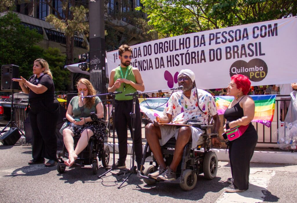 Foto colorida da Parada do Orgulho PCD em Salvador. Da esquerda para a direita aparecem uma mulher branca, de roupa preta, fazendo interpretação de Libras. Uma mulher branca usuária de cadeira de rodas, um homem branco de pé ao seu lado. Outro homem usuário de cadeira de rodas, preto e com roupas brancas. E no canto direito, uma mulher com nanismo branca de cabelo rosa e usando roupas pretas. Ao fundo, faixa branca com os logos do ValePCD e QuilomboPCD e a frase: "1° Parada do Orgulho da Pessoa com Deficiência da História do Brasil".