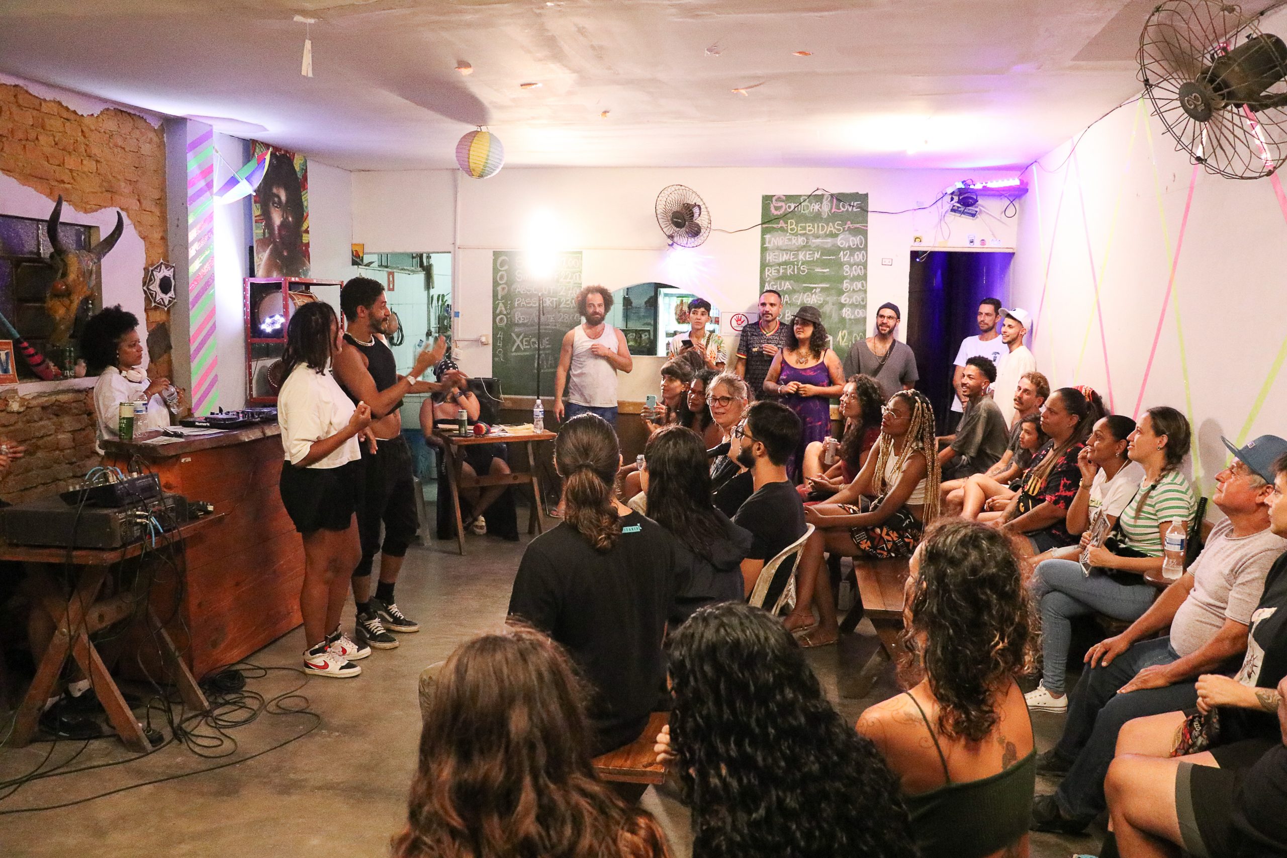 Foto colorida de evento do Slam do Corpo em Ubatuba. No canto esquerdo da imagem, Leo Castilho e Erika Mota estão de pé e olham para as pessoas na plateia, que estão sentadas. Ao fundo, algumas pessoas de pé assistem a apresentação.