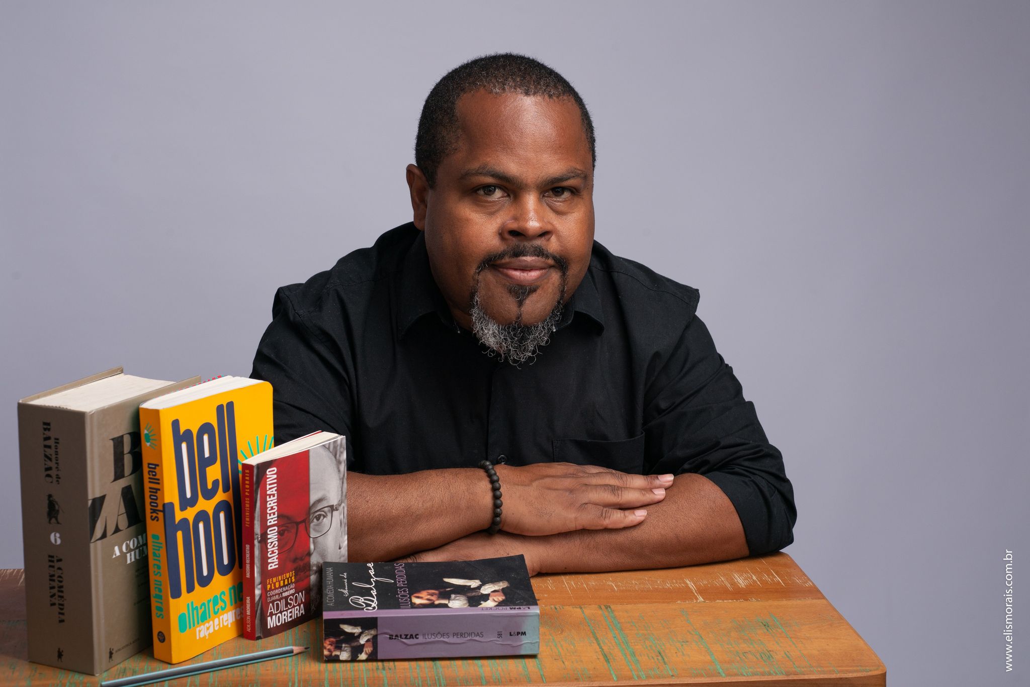 Foto colorida do doutor em História João Heitor. João é negro e usa uma camisa preta, ele está sentado com os braços cruzados sobre uma mesa onde estão alguns livros. Ao fundo, parede cinza