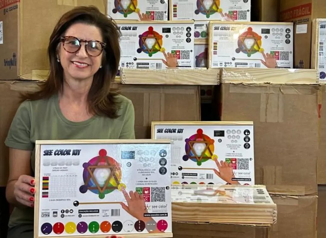 Foto colorida de Sandra Marchi com kit pedagógico do See Color. Sandra é uma mulher branca com cabelo castanho e usa um óculos vermelho escuro. Ela sorri, enquanto segura uma caixa branca com detalhes coloridos e o desenho dos materiais do See Color