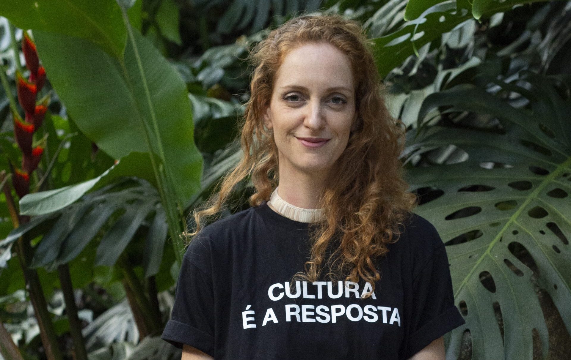 Foto colorida de Laila Zaid. Ela é uma mulher branca com cabelos ruivos. Ela sorri e olha para a foto. Ela veste uma camiseta preta com a frase "Cultura é a resposta". Ao fundo, arbustos e plantas verdes