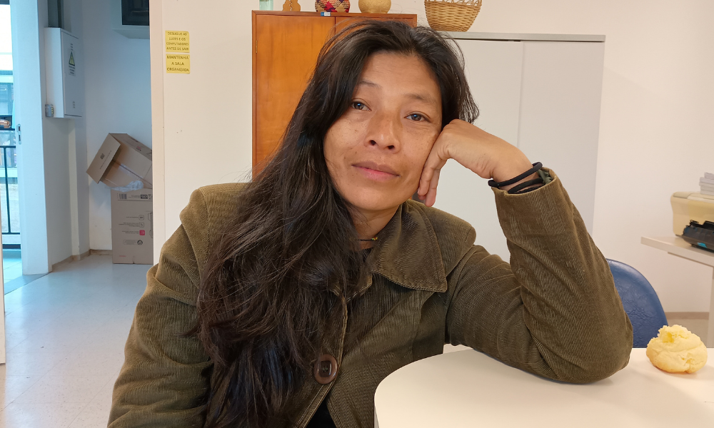 Foto colorida de Patrícia Ferreira Ixapy. Ela é uma mulher indígena guarani de cabelos pretos, lisos e longos. Ela usa um casaco marrom e está com o braço esquerdo apoiado em uma mesa. Ao fundo, armário e parede branca