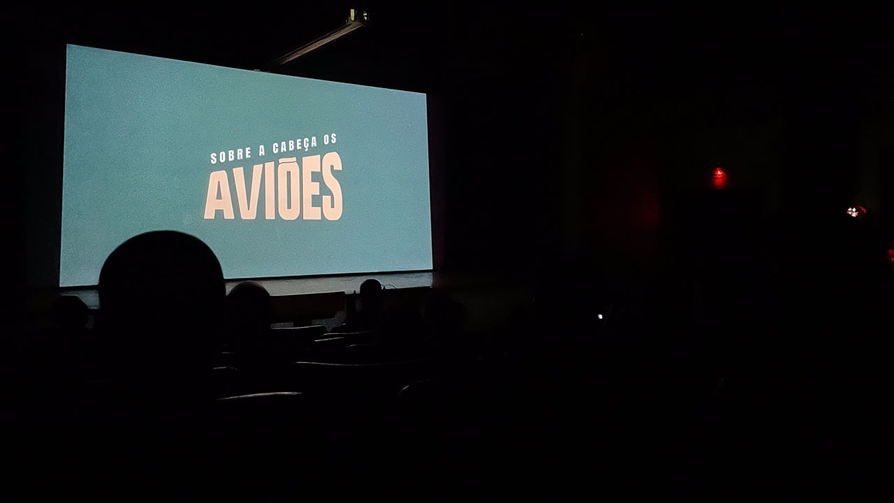 Foto colorida de sala de cinema com exibição do curta "Sobre a cabeça, os aviões"