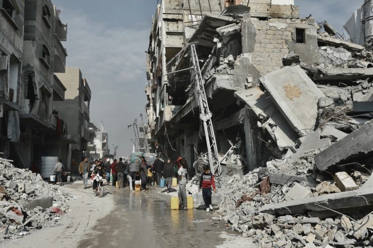Foto colorida de pessoas buscando água ao lado de um prédio que corre o risco de desabar em Beit Lahia, no Norte de Gaza.