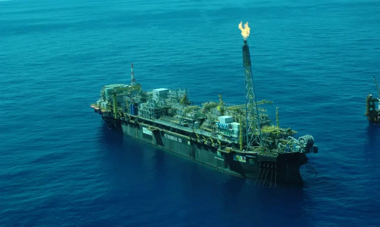 Foto colorida de plataforma de petróleo operando no mar. Em uma das torres, é possível ver uma chama da queima de combustíveis