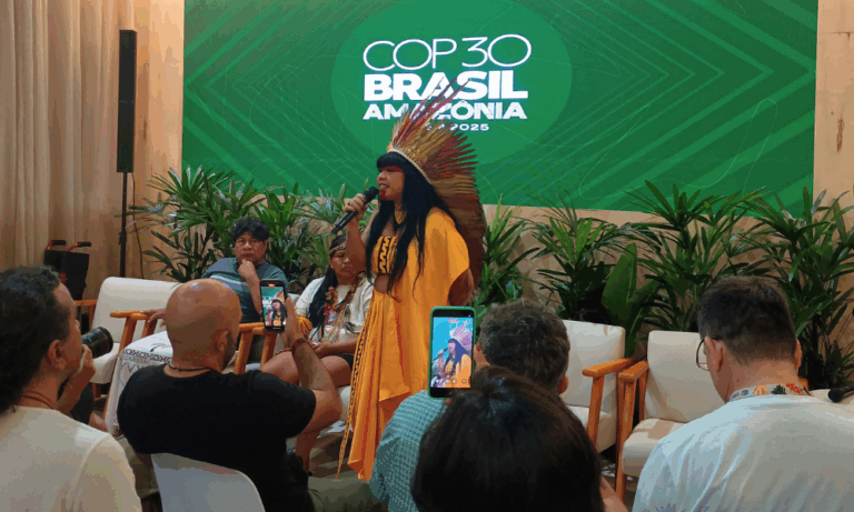 Foto colorida da deputada Célia Xakriabá. Ela está usando um vestido amarelo e um cocar amarelo com vermelho. Ela é uma mulher indígena com cabelos pretos e segura um microfone. Ao fundo, painel verde com logo da COP30