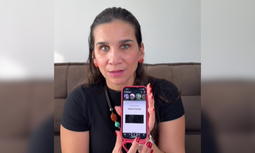 Captura de tela colorida com foto de Geisa Farini. Ela é uma mulher branca com cabelos pretos. Ela está segurando um celular com o Instagram aberto e com a tela voltada para frente. Ela usa um colar com contas coloridas e uma camiseta preta. Ao fundo, sofá marrom e parede branca