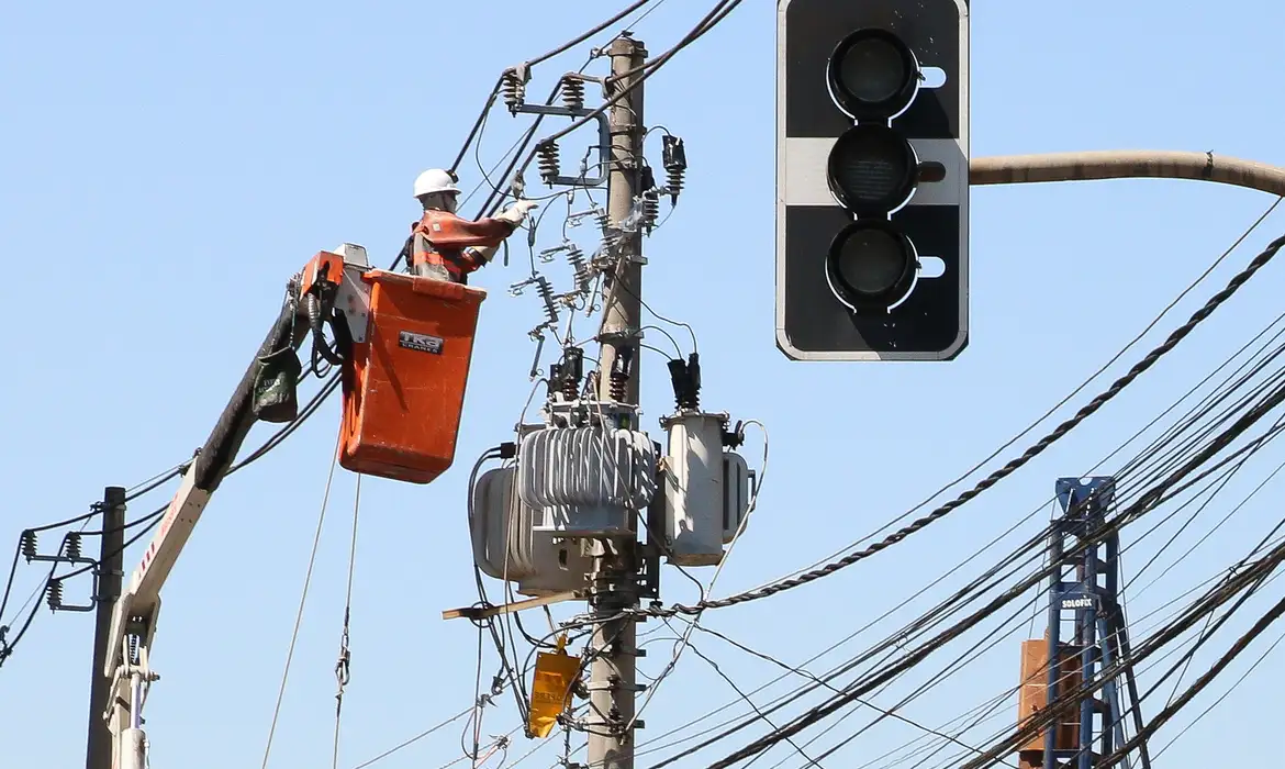 Foto colorida de trabalhador do setor elétrico em cesto, trabalhando em linha de rede elétrica, ao fundo, céu azul