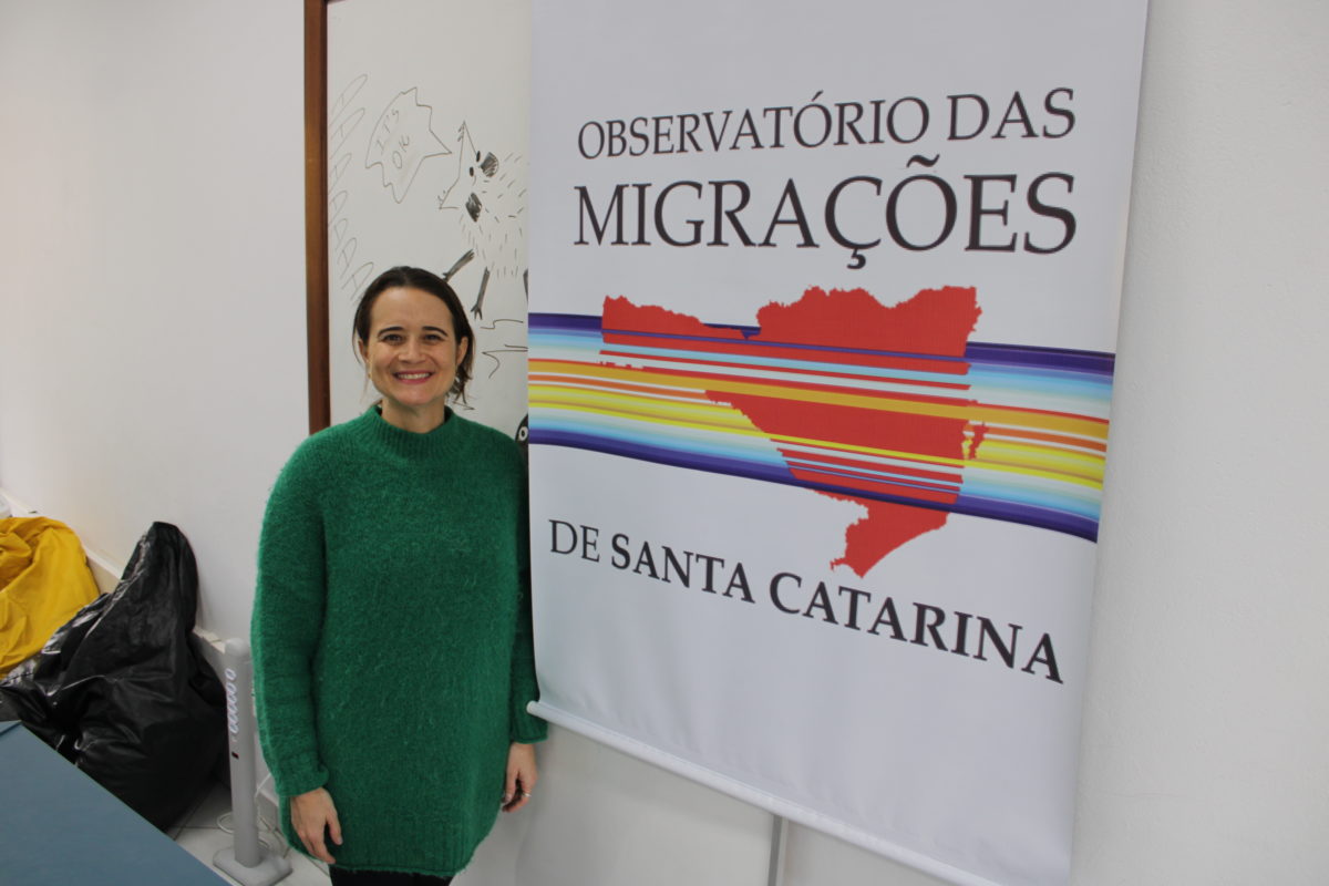 Foto colorida de Lina Magalhães, uma mulher branca de cabelo preto e que usa um agasalho verde ao lado do banner do Observatório de Migrações de Santa Catarina
