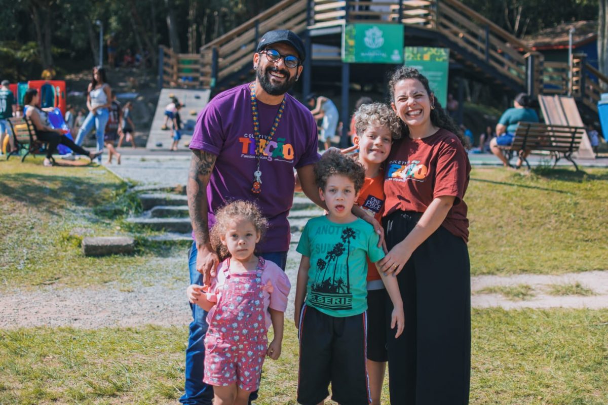 Foto colorida de Thiago com sua esposa e seus três filhos. Eles posam para a foto e sorriem. Ao fundo, um parque